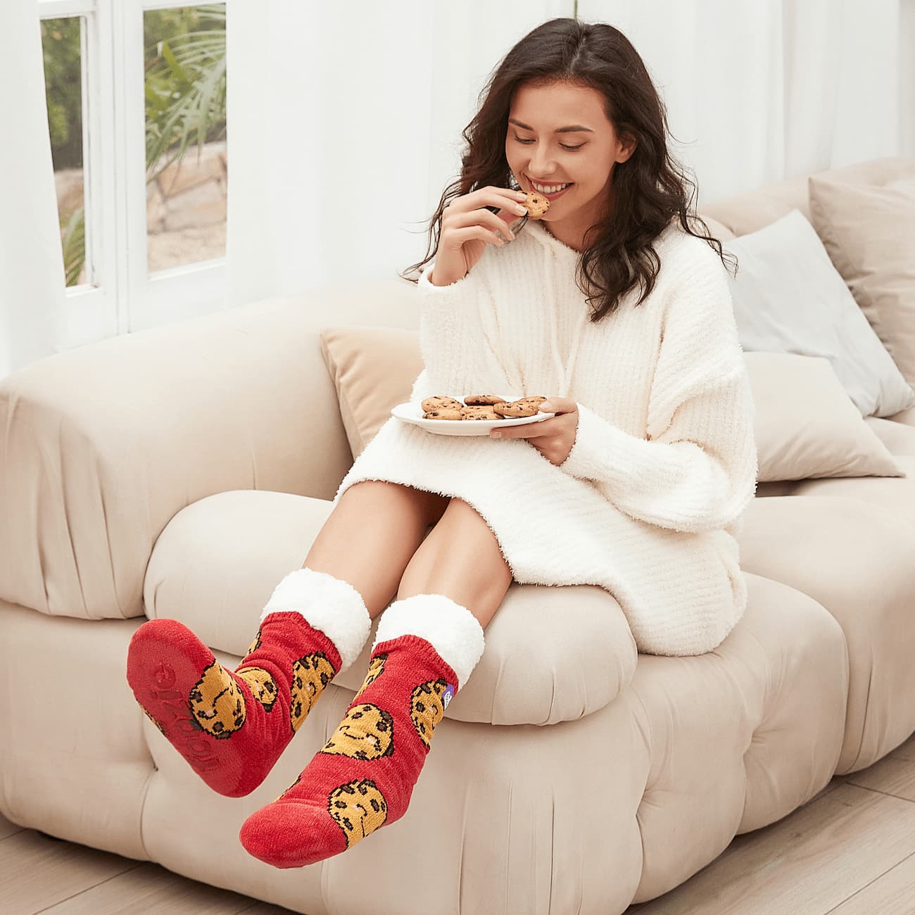 The Foozie™ Cookie Red slipper socks, worn by a woman eating a cookie on the sofa with a plate of cookies